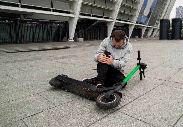 Man fallen from scooter holding his knee and feeling pain. Green electric scooter lying on asphalt. Stylish man in gray hoody sits on the ground and has knee pain. Eco-friendly transport concept.