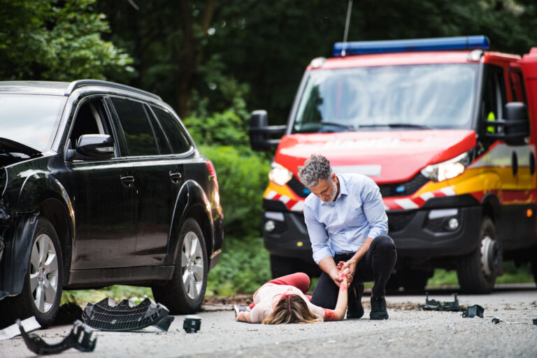 A man helping a young woman lying unconscious on the road after a car accident.